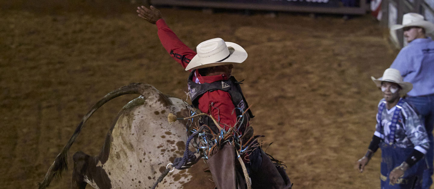 Cowboy riding a bull at a rodeo wearing a red shirt.